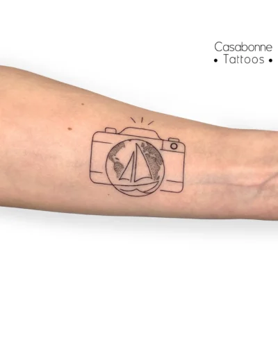 Tatouage ligne fine d'un appareil photo avec un voilier dans l'objectif, sur l'avant-bras, à Toulouse