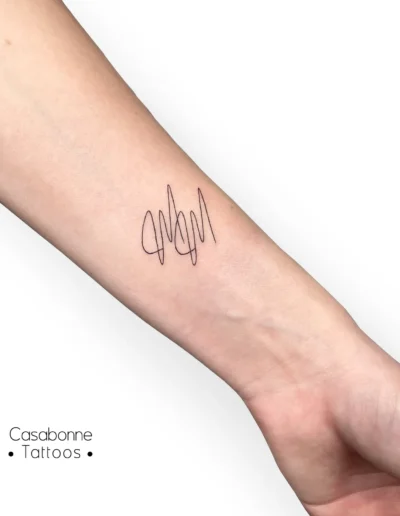 Tatouage ligne fine de la lettre 'M' en écriture stylisée, sur l'avant-bras, à Toulouse