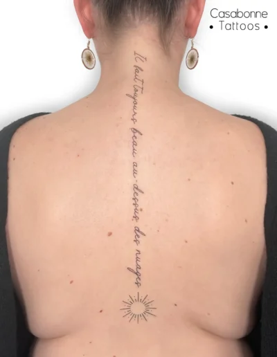 Tatouage ligne fine d'une phrase le long de la colonne, se terminant par un soleil, sur le dos, à Toulouse