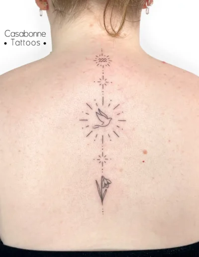 Tatouage ligne fine composé d'un signe astrologique, d'un oiseau et d'une fleur de perce-neige, le long de la colonne, à Toulouse
